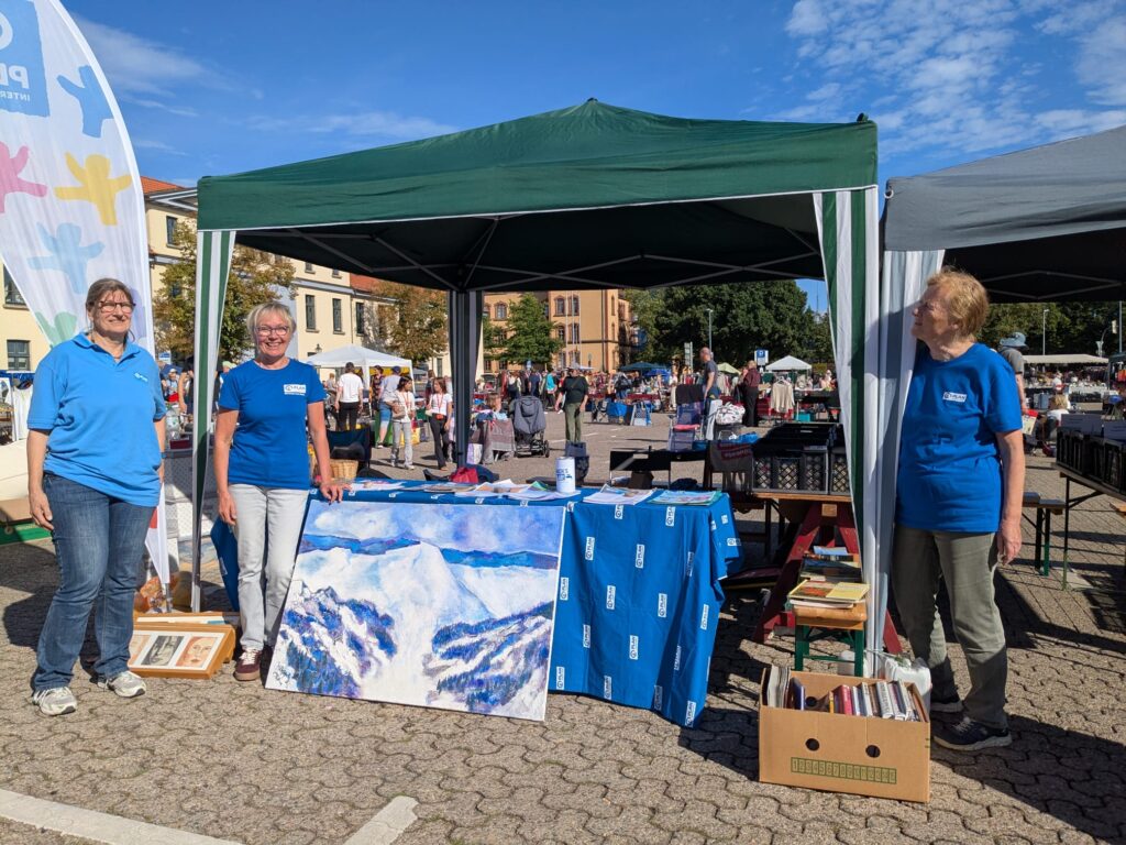 Drei Frauen stehen neben einem Marktstand unter einem grünen Vordach und präsentieren Bücher sowie ein großes Gemälde mit Bergmotiven. In der sonnigen Kulisse im Freien sind weitere Stände und Menschen zu sehen.