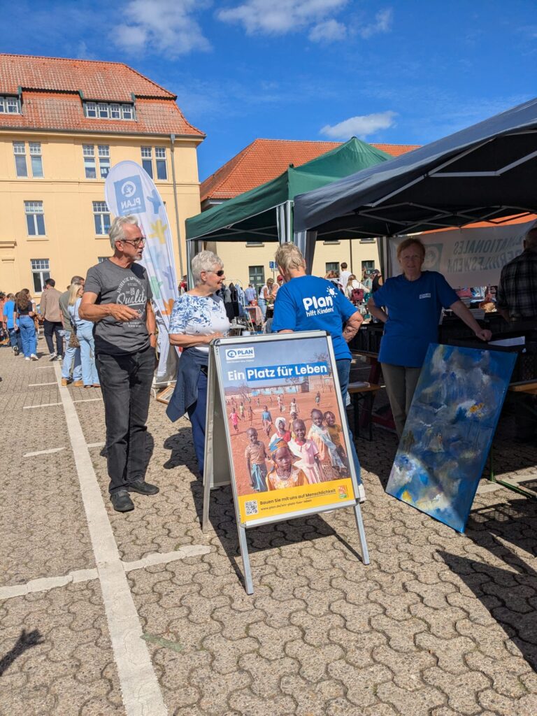 Bei einer Veranstaltung im Freien mit Ständen und Zelten stehen Menschen beisammen und unterhalten sich. Im Vordergrund ist ein blaues Schild mit der Aufschrift „Ein Platz für Leben“ und Fotos von Kindern zu sehen. Es ist ein strahlender, sonniger Tag mit blauem Himmel und einem gelben Gebäude im Hintergrund.