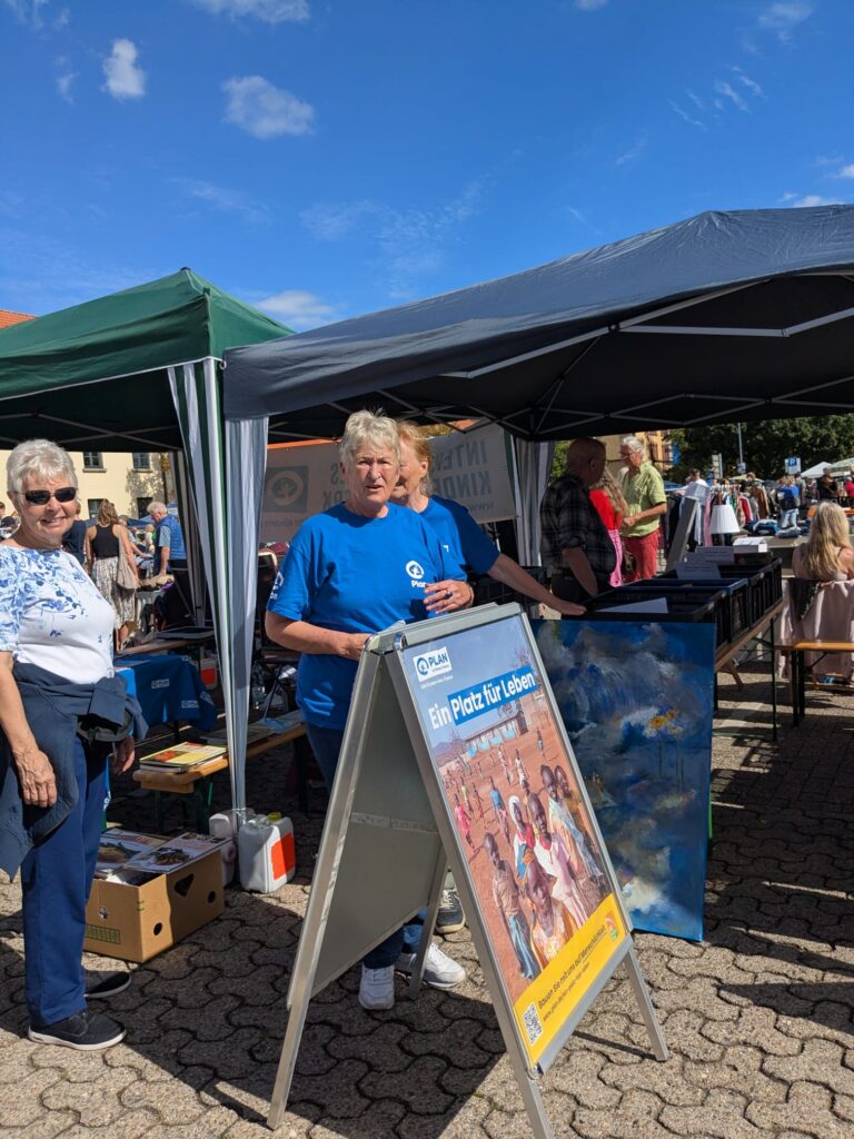 Eine Frau in einem blauen Hemd steht an einem Informationstafel unter einem Vordach auf einem Straßenfest, im Hintergrund sind an einem sonnigen Tag weitere Menschen und Marktstände zu sehen.