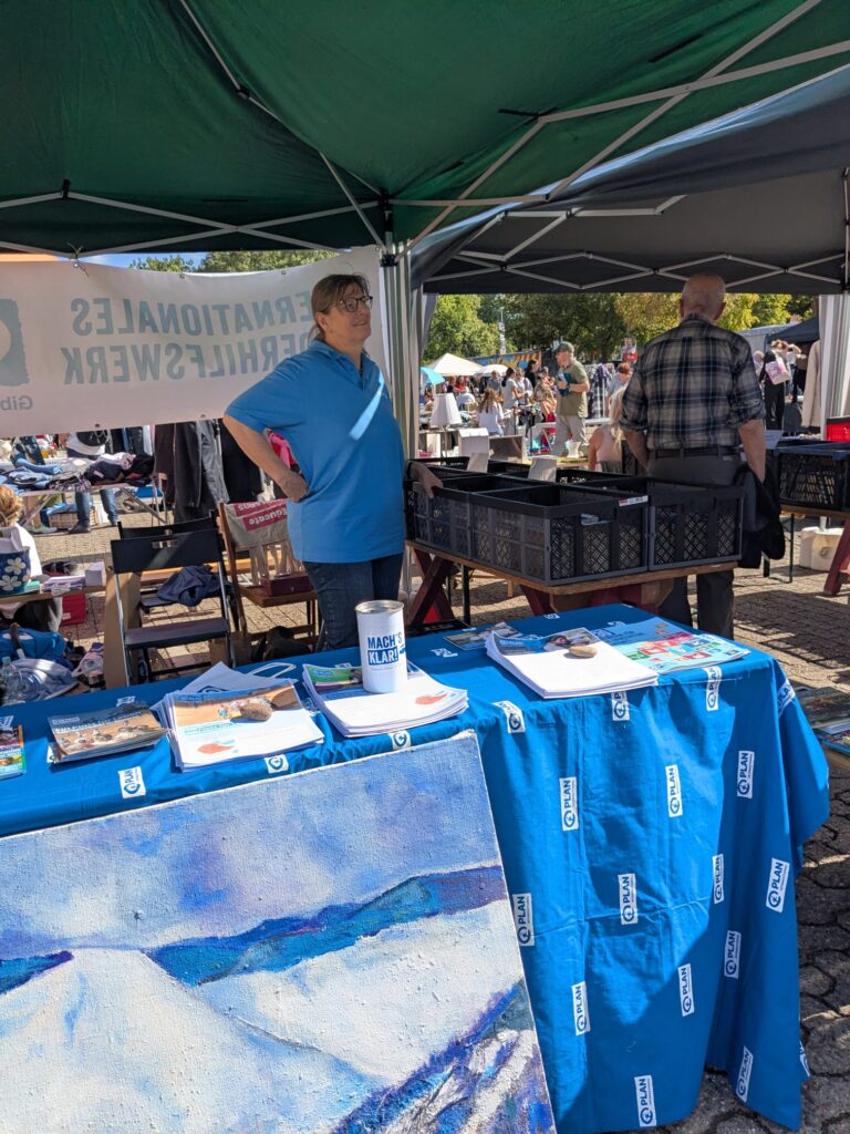 Auf einem Markt im Freien steht eine Person hinter einem Tisch, der mit Büchern und Kunstwerken bedeckt ist. Eine weitere Person steht in der Nähe. Im Vordergrund sind eine blaue Tischdecke und ein großes Gemälde zu sehen. Im Hintergrund sind weitere Menschen und Stände zu erkennen.