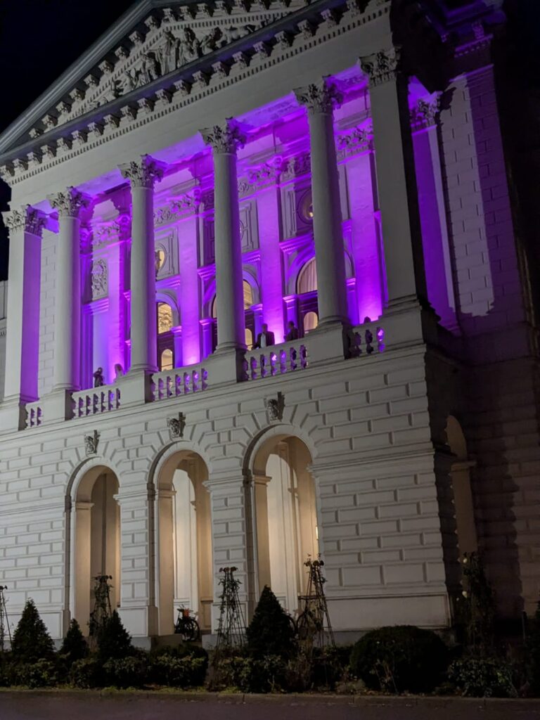 Ein neoklassizistisches Gebäude, dessen obere Säulen und Bögen nachts in leuchtendem Violett erstrahlen.
