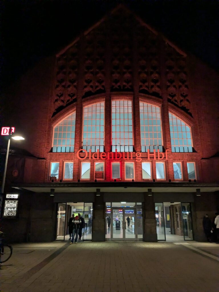 Der Oldenburger Hauptbahnhof bei Nacht, mit einer großen Backsteinfassade, beleuchteten Fenstern und einem leuchtend roten Schild.